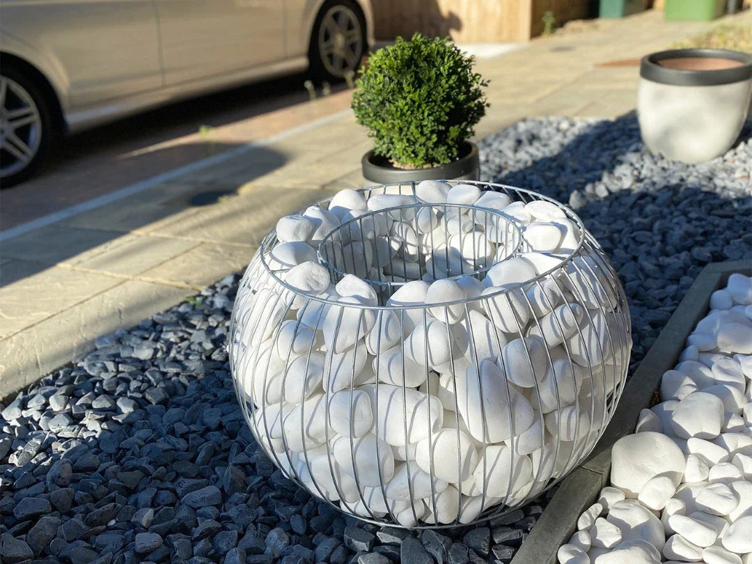 Round gabion basket filled with white stones on a bed of dark gravel, next to a small potted shrub and parked car.