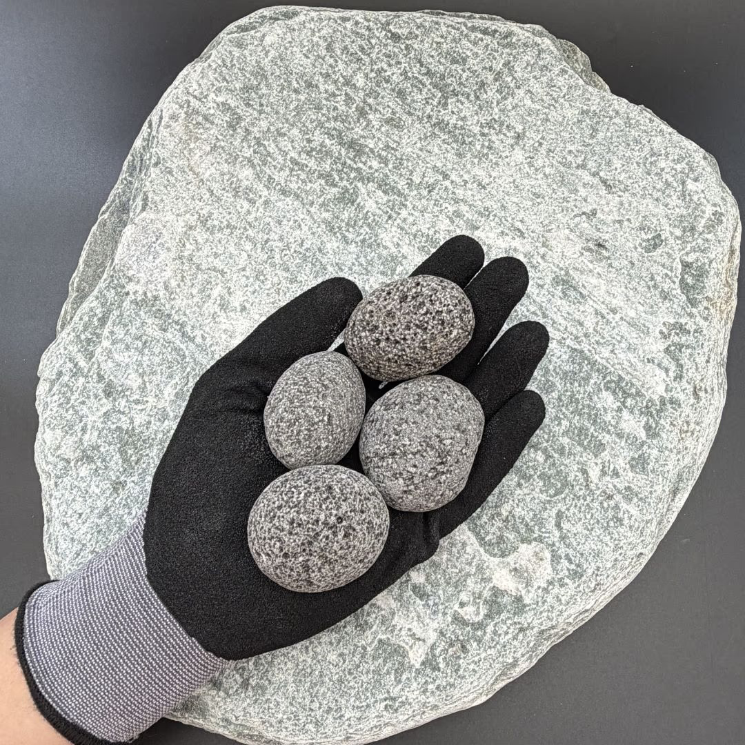 Gloved hand holding four black lava volcanic stones, 30-50mm, on a large textured rock background.