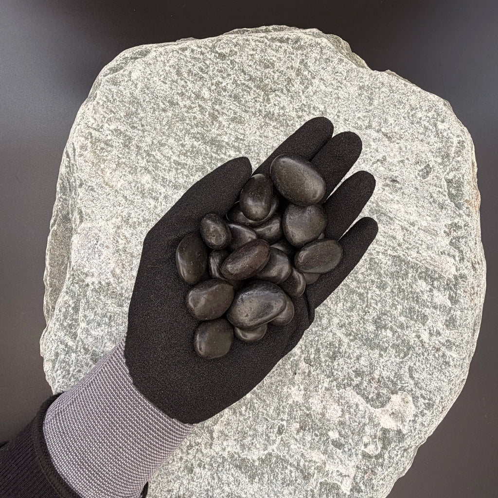 Gloved hand holding small polished black pebbles of size 10 to 30mm over large grey stone surface.