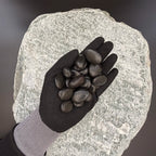 Gloved hand holding small polished black pebbles of size 10 to 30mm over large grey stone surface.