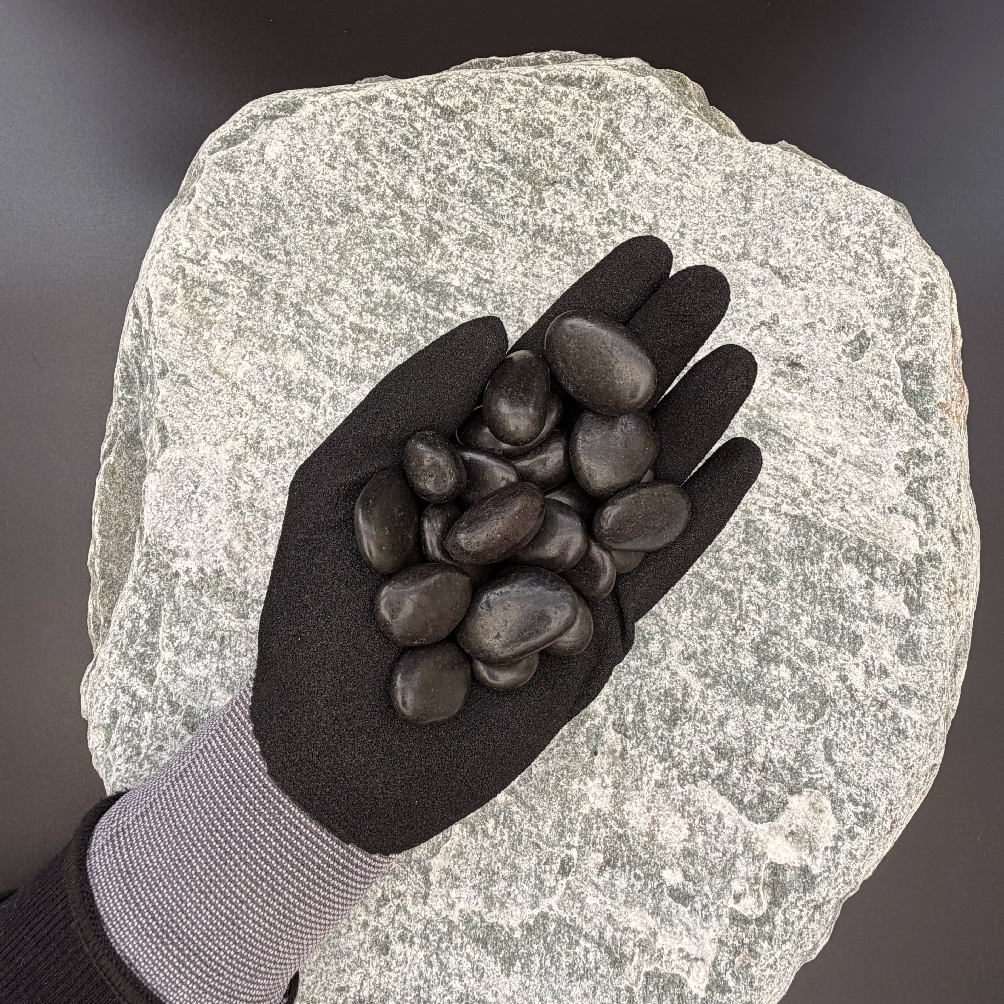 Gloved hand holding small polished black pebbles of size 10 to 30mm over large grey stone surface.