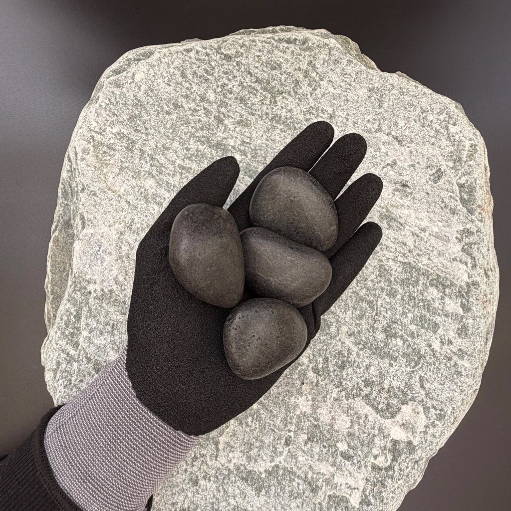 Gloved hand holding medium polished black pebbles of size 30 to 50mm over large grey stone surface.