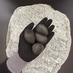 Gloved hand holding medium polished black pebbles of size 30 to 50mm over large grey stone surface.