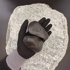 Gloved hand holding large polished black pebbles of size 50 to 80mm over large grey stone surface.