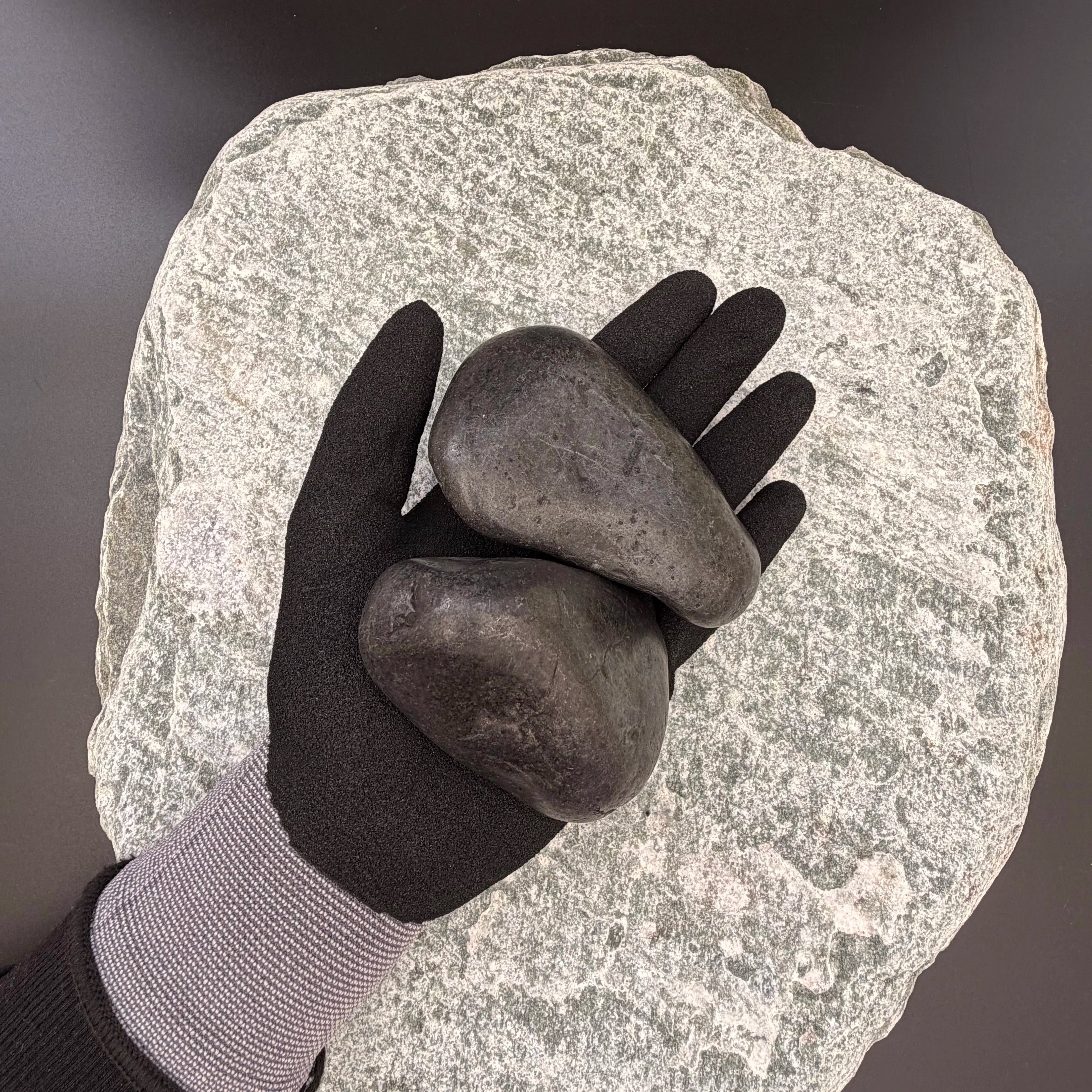 Gloved hand holding large polished black pebbles of size 50 to 80mm over large grey stone surface.