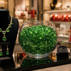 Vase filled with green glass stones beside jewelry display with necklace, earrings, and ring on black stands.