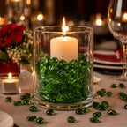 Candle in a glass vase with green decorative stones on a table setting, surrounded by red flowers and glassware.