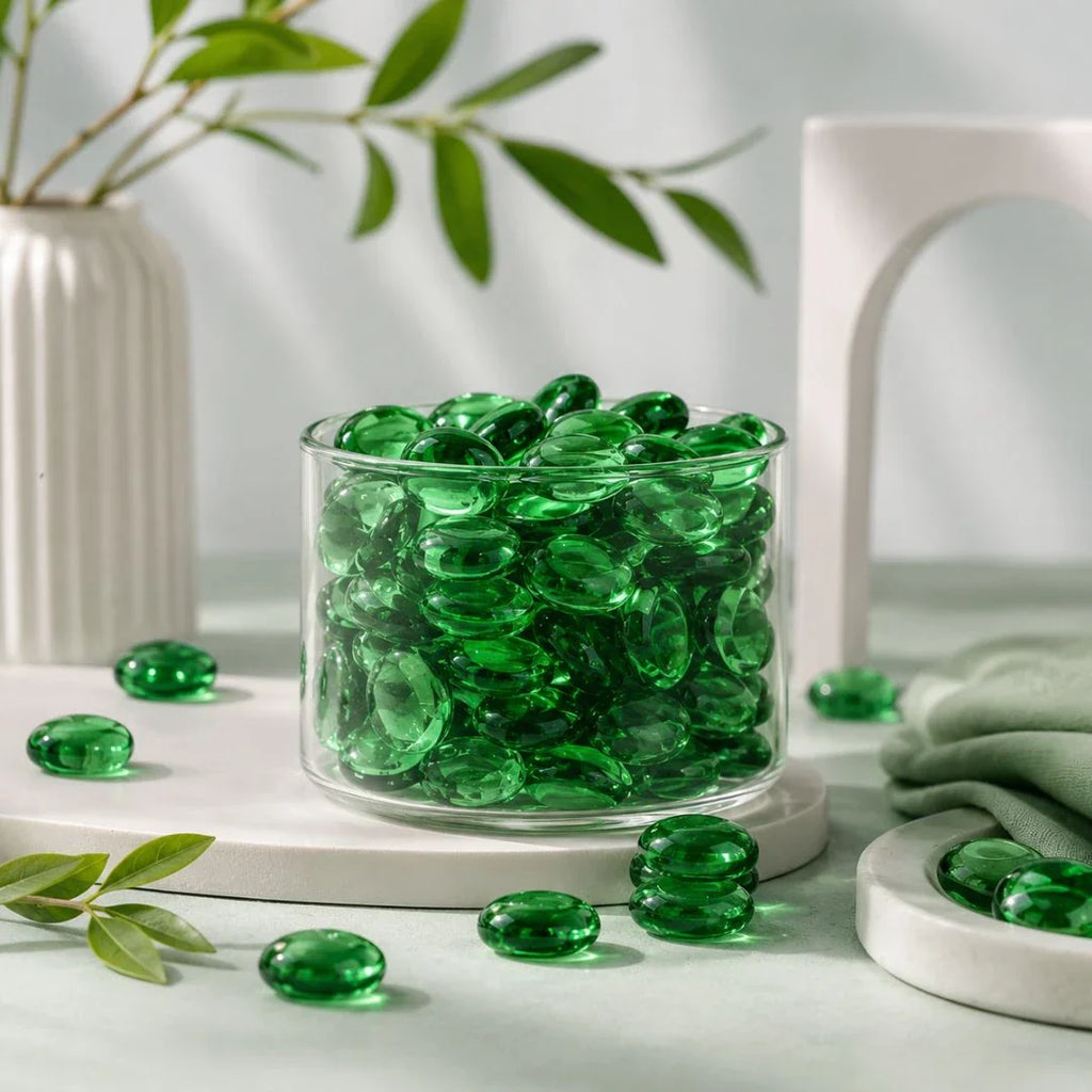 Green decorative glass stones in a clear vase, with scattered stones and greenery on a white surface.