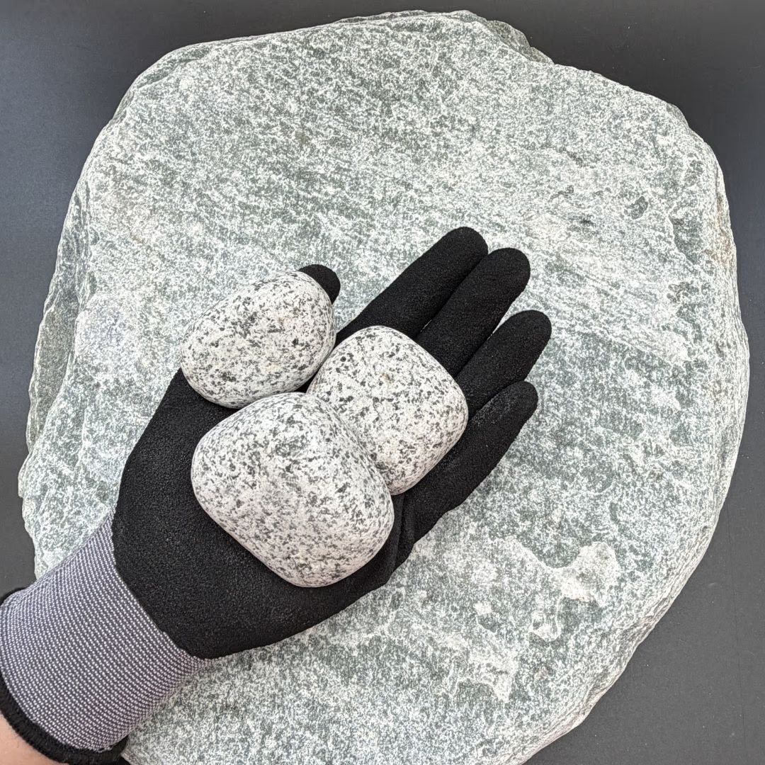 Gloved hand holding three grey granite pebbles on a larger stone background.
