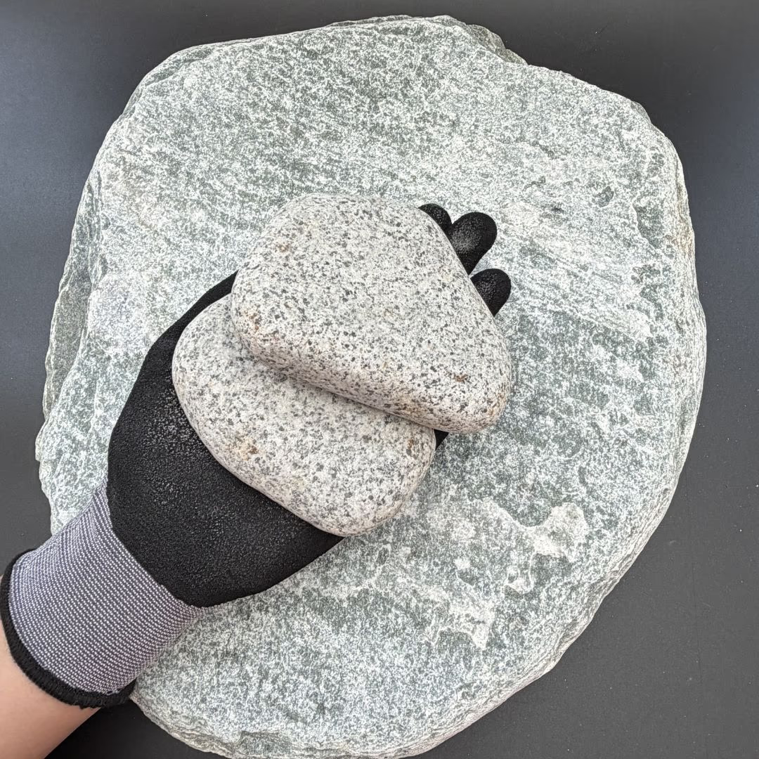 Gloved hand holding two grey granite pebbles on a larger stone background.