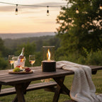 Outdoor picnic table with black tabletop fireplace, wine, glasses, cheese, grapes, and a blanket at sunset.