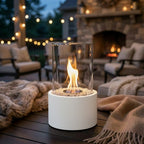 White tabletop bioethanol fireplace with glass cylinder, set on a patio table, surrounded by cozy seating and string lights.