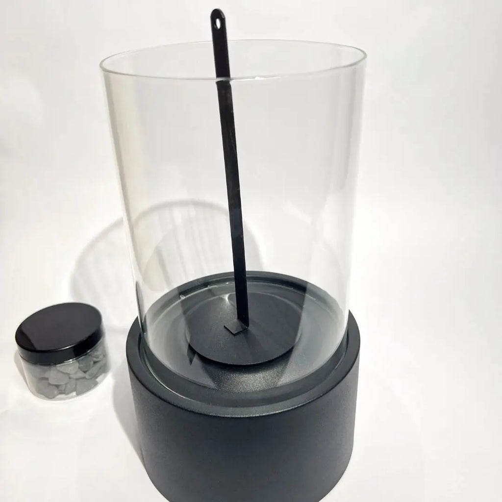 Helios tabletop fireplace with glass cylinder and metal base, includes fire extinguisher tool, jar of stones nearby.