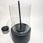 Helios tabletop fireplace with glass cylinder and metal base, includes fire extinguisher tool, jar of stones nearby.
