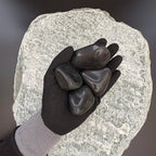 Gloved hand holding two medium high-gloss polished black pebbles on a large textured stone background.