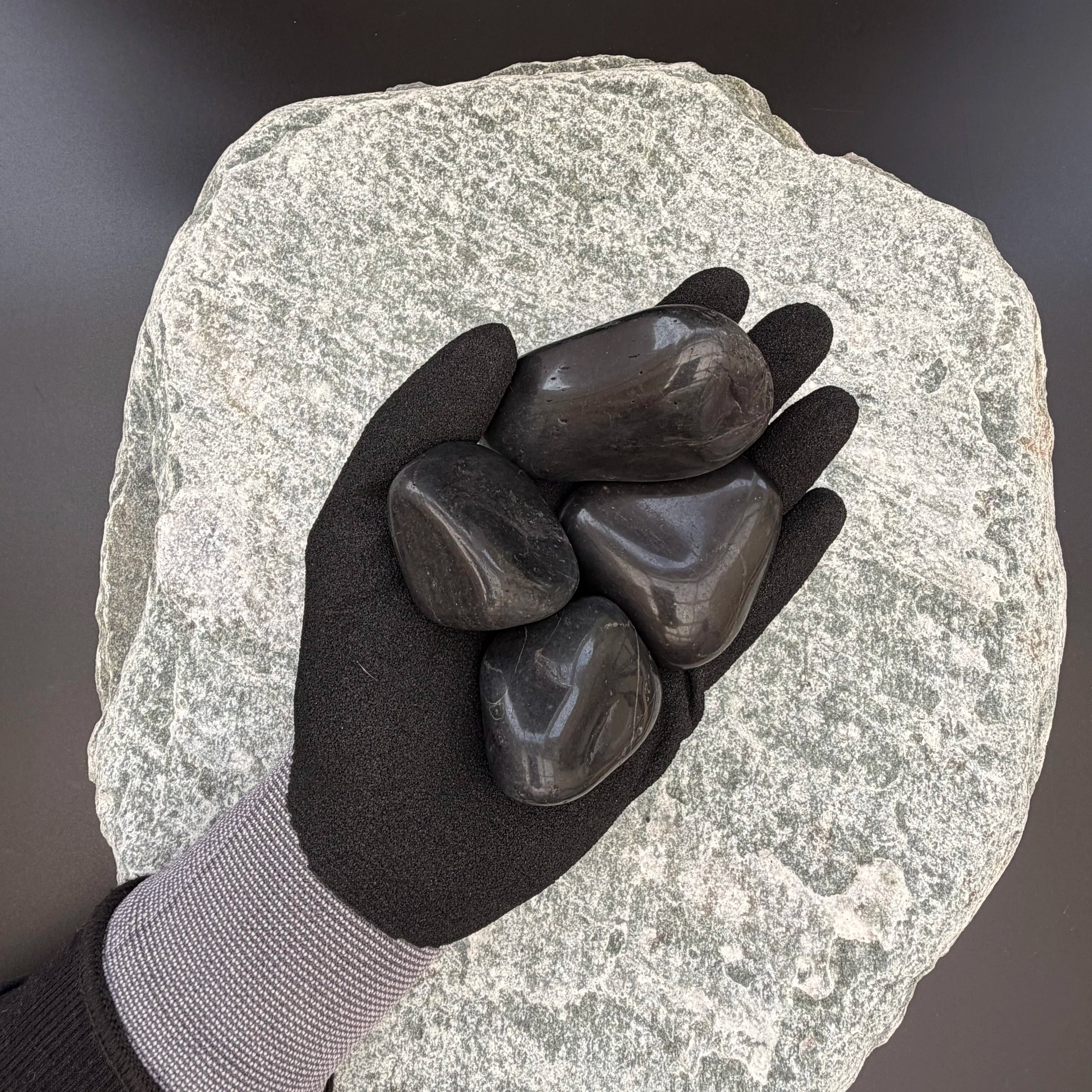Gloved hand holding two medium high-gloss polished black pebbles on a large textured stone background.