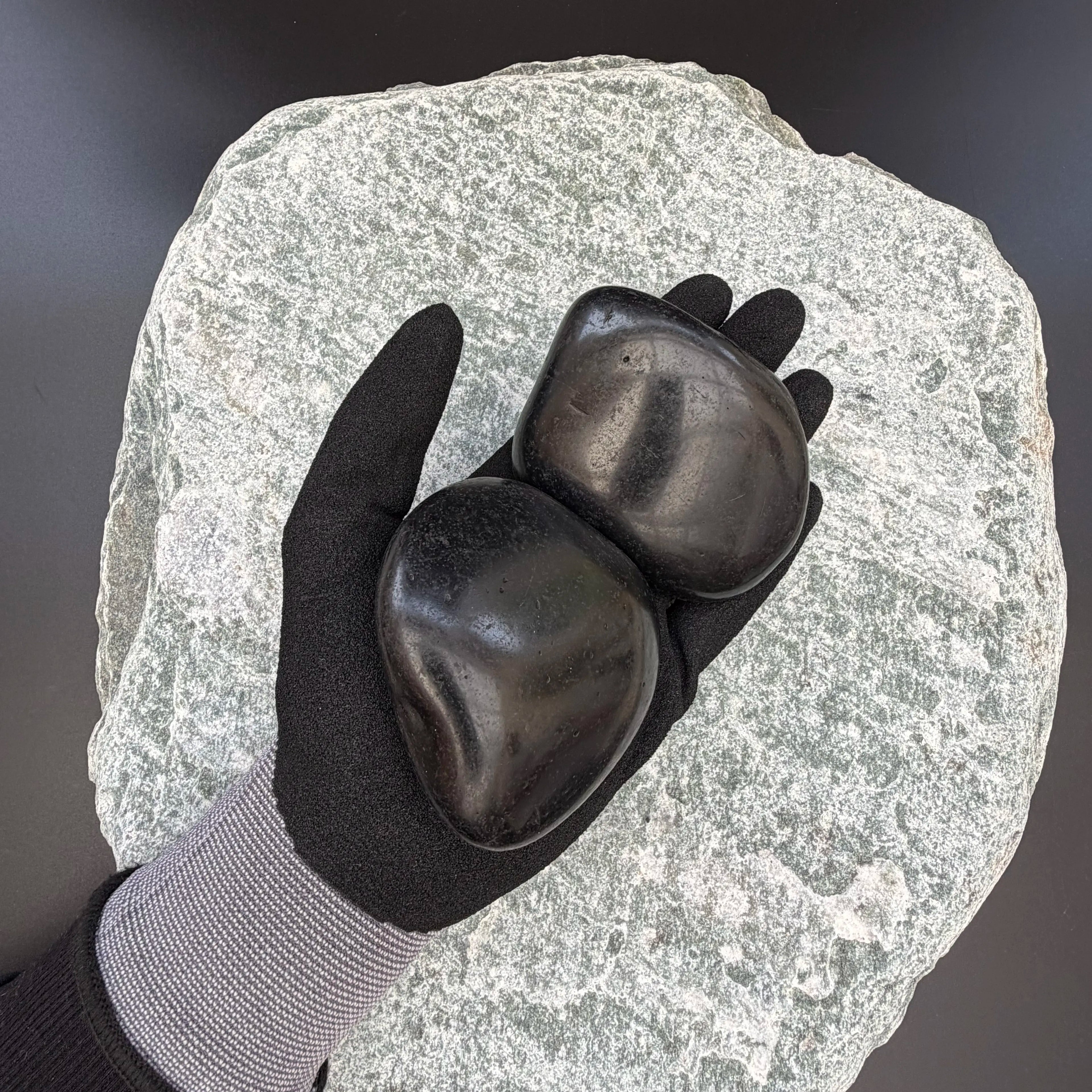 Gloved hand holding two large high-gloss polished black pebbles on a large textured stone background.