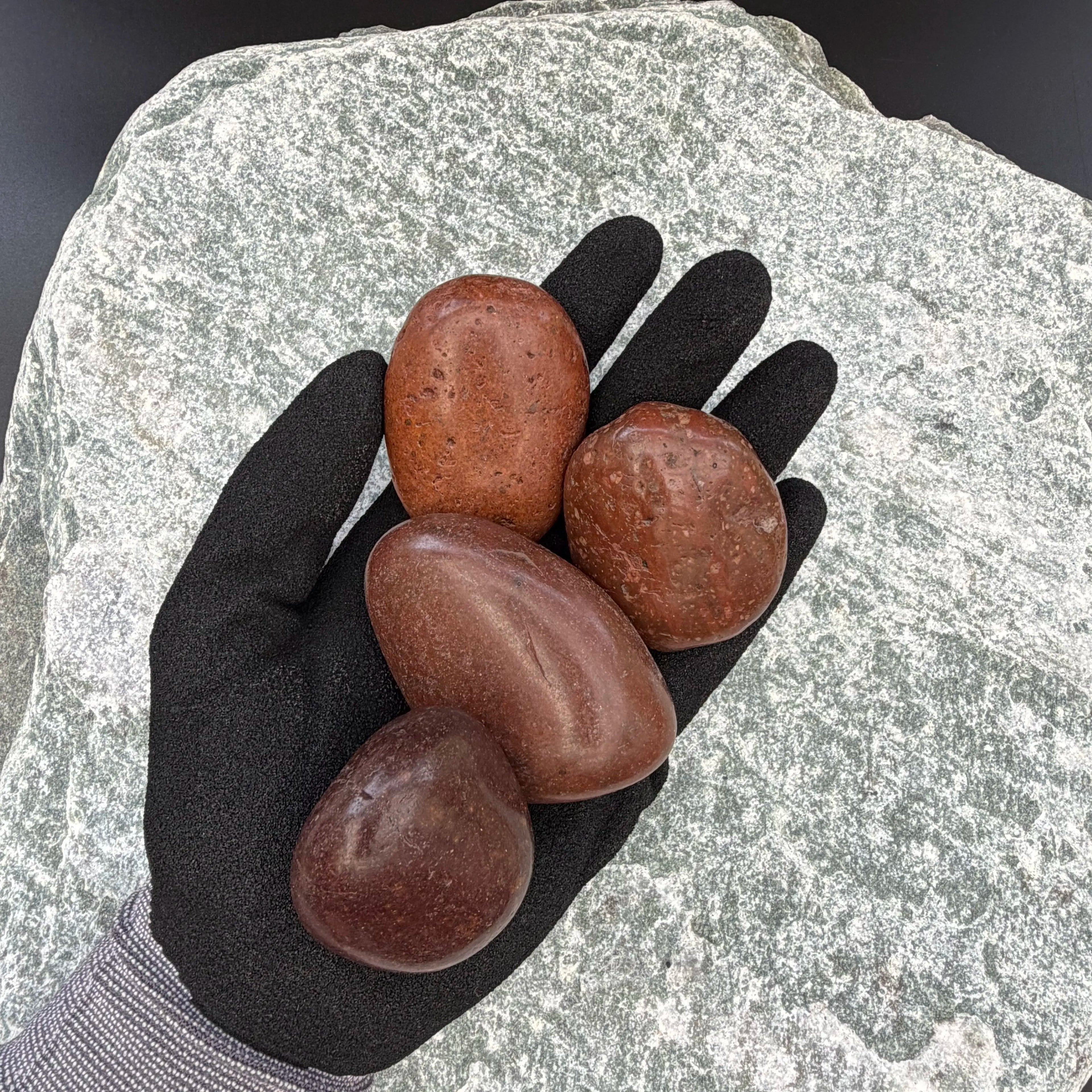 Hand in black glove holding four polished cherry mahogany pebbles on textured stone background.