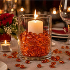 Vase with orange glass stones and lit candle on a table, surrounded by scattered stones and a floral arrangement.