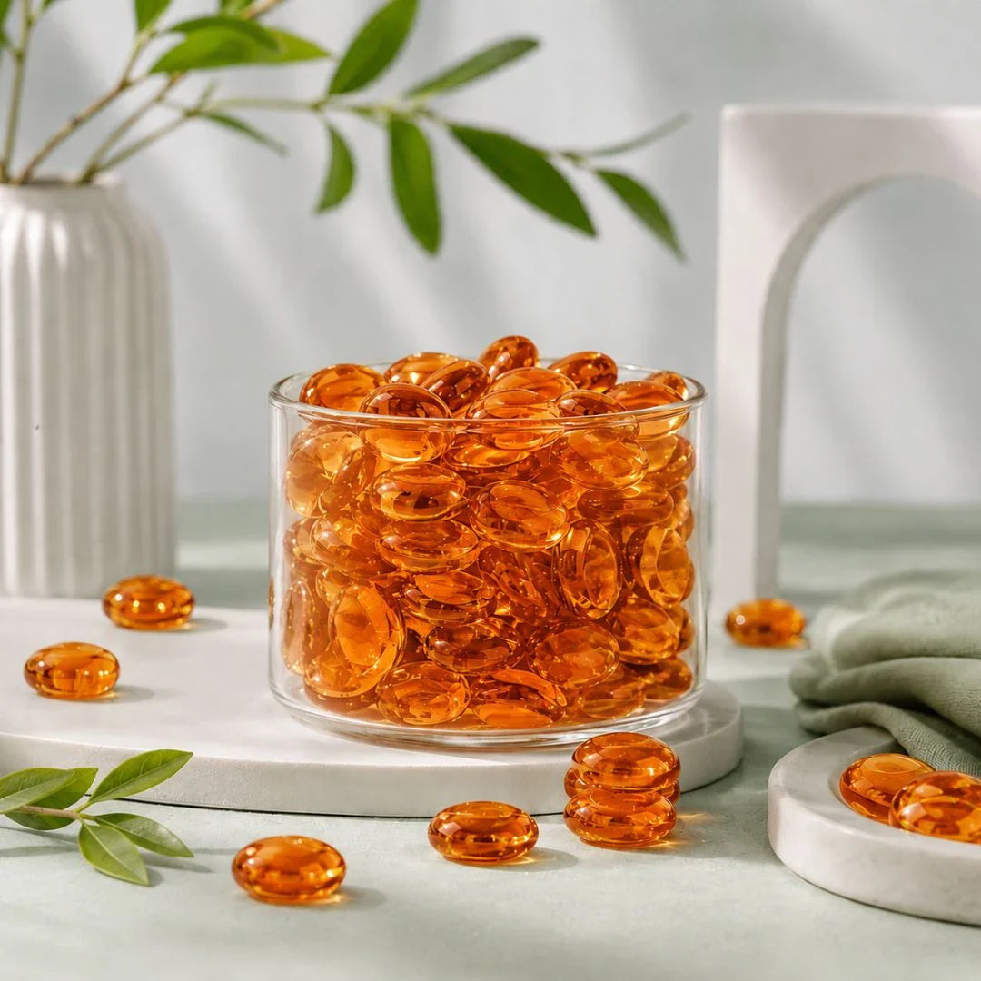 Orange glass stones in a clear vase on a table with green leaves and white decor.