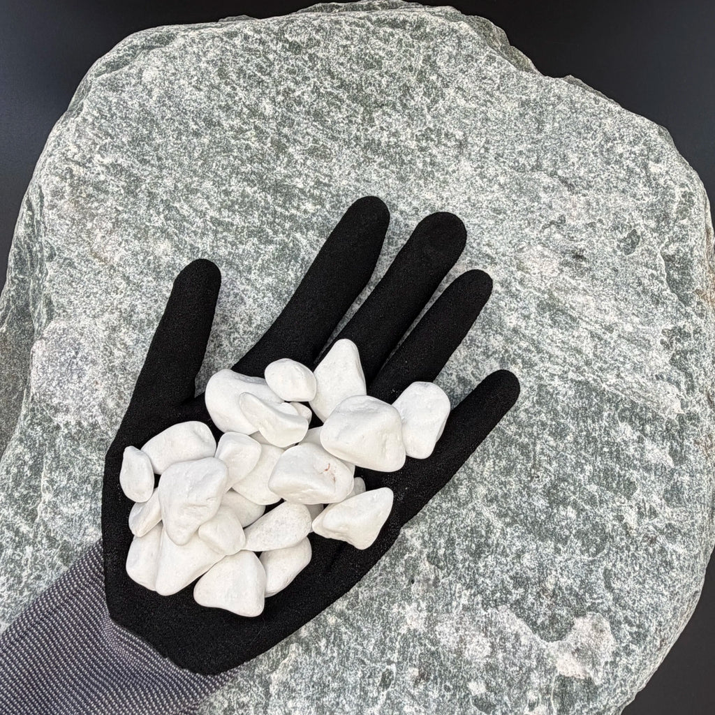 Black-gloved hand holding a small, smooth snow white thassos marble pebble on a textured stone background.
