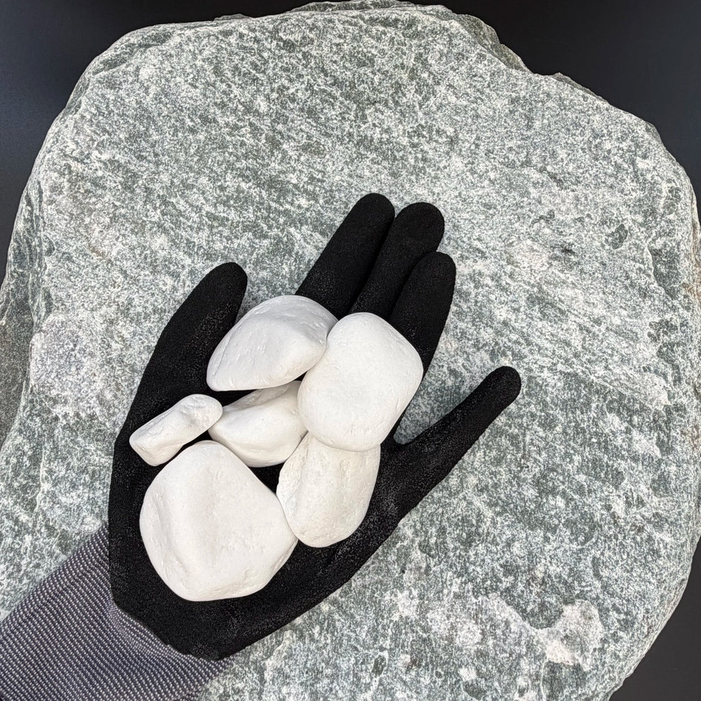 Black-gloved hand holding a medium, smooth snow white thassos marble pebble on a textured stone background.