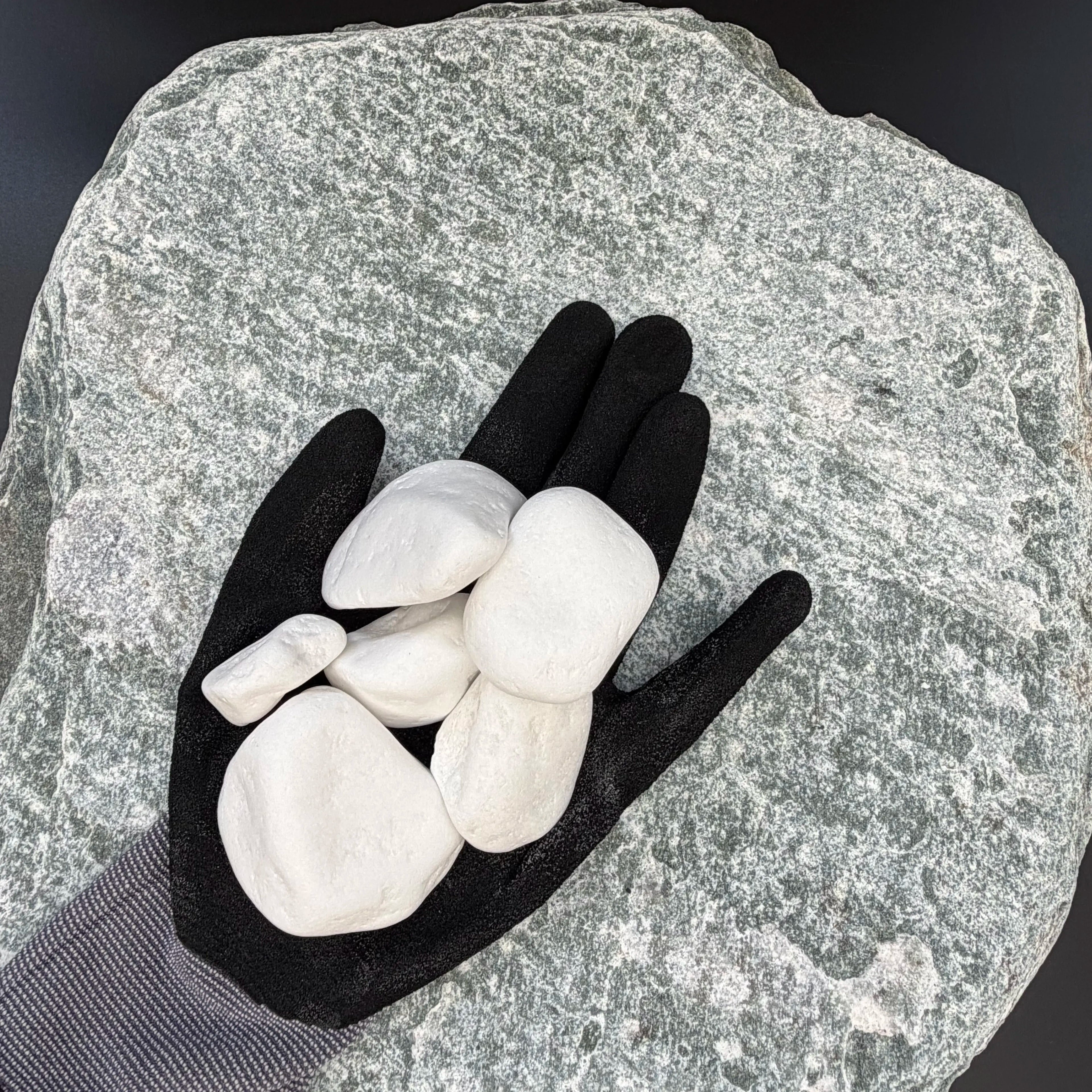 Black-gloved hand holding a medium, smooth snow white thassos marble pebble on a textured stone background.
