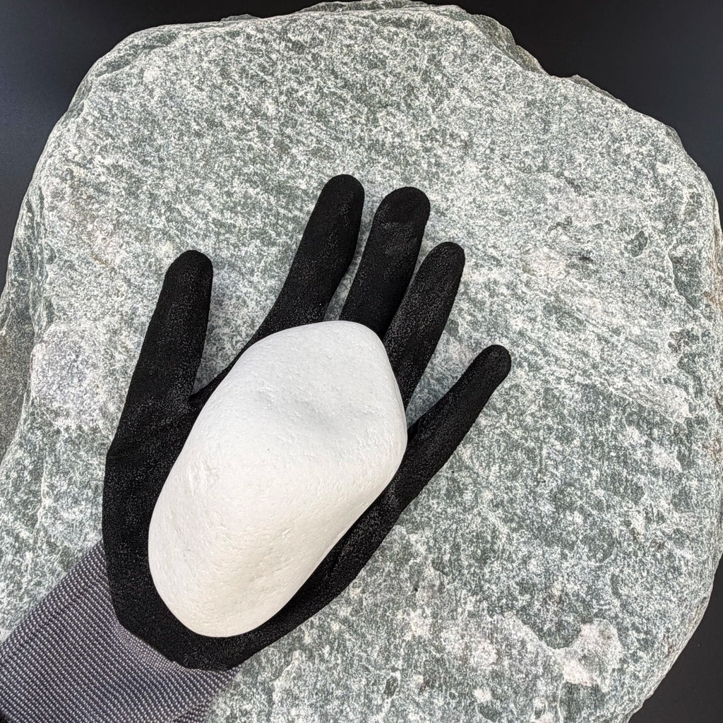 Black-gloved hand holding a large, smooth snow white thassos marble pebble on a textured stone background.