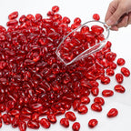 Hand scooping glossy red glass pebbles with a clear scoop on a white surface.