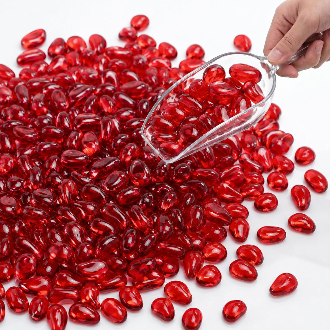 Hand scooping glossy red glass pebbles with a clear scoop on a white surface.