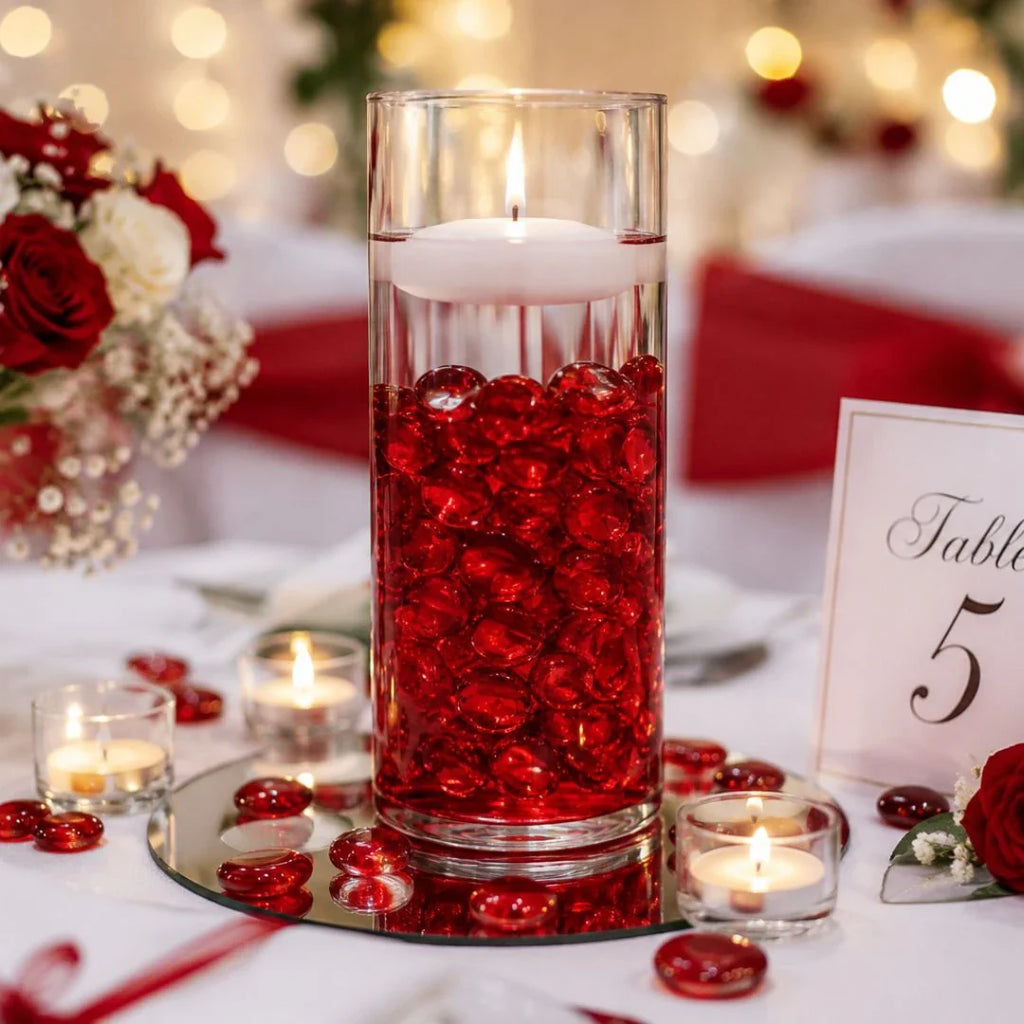 Vase with red glass stones and floating candle, surrounded by flowers and tealights on wedding table.