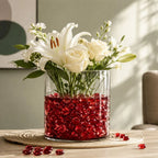 Clear vase with white flowers and red glass stones on a woven mat, sunlight casting shadows on the wall.