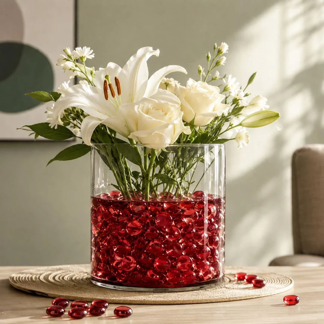 Clear vase with white flowers and red glass stones on a woven mat, sunlight casting shadows on the wall.
