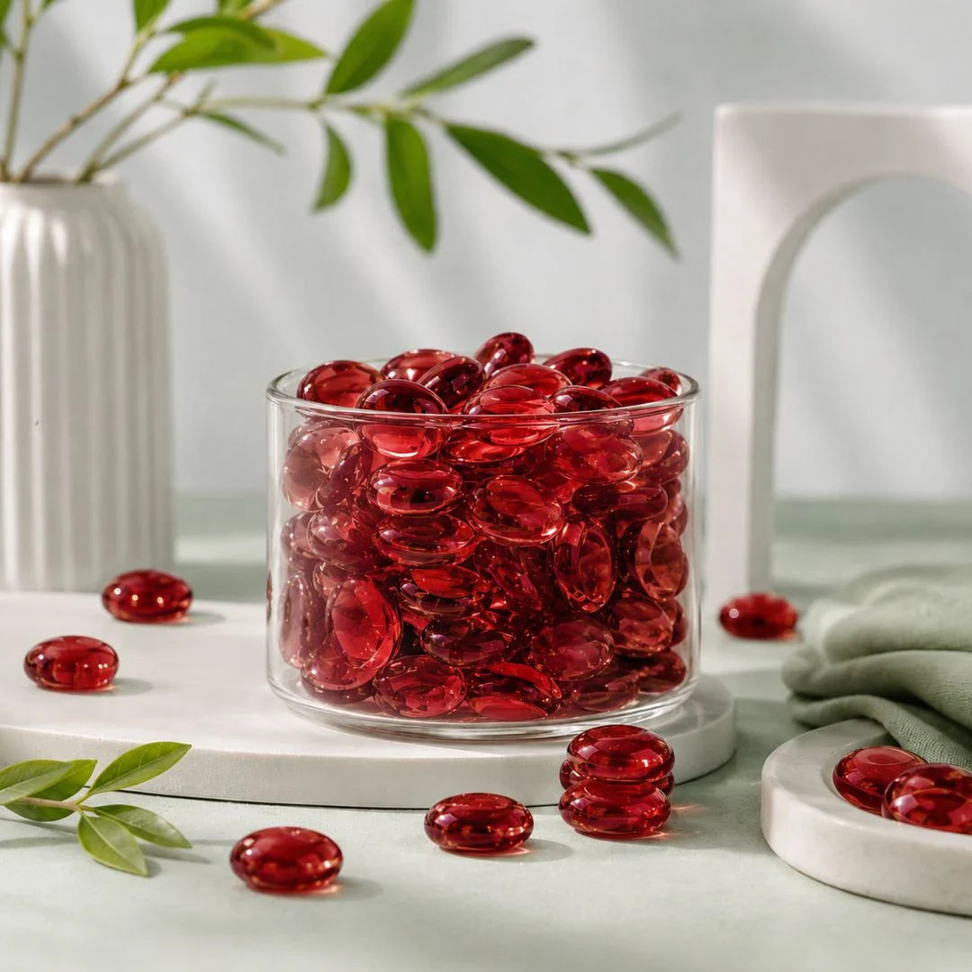Clear vase filled with red glass stones on a table with scattered stones, a white vase, and greenery.