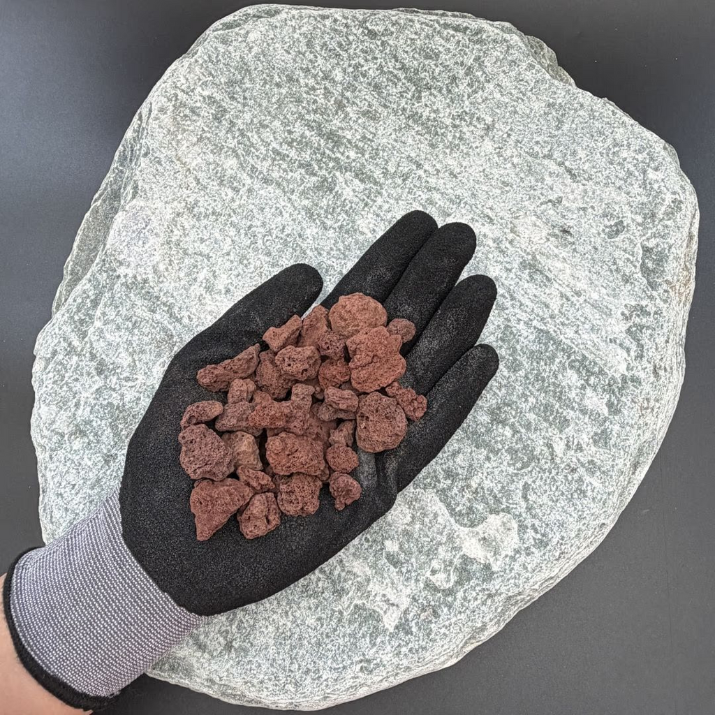 Gloved hand holding red lava volcanic gravel rocks on a large gray stone background.