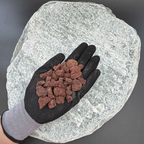 Gloved hand holding red lava volcanic gravel rocks on a large gray stone background.