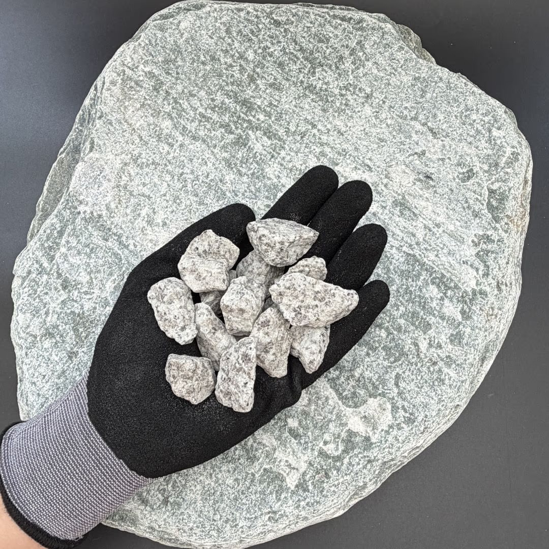 Gloved hand holding several pieces of silver granite gravel on a large flat stone background.