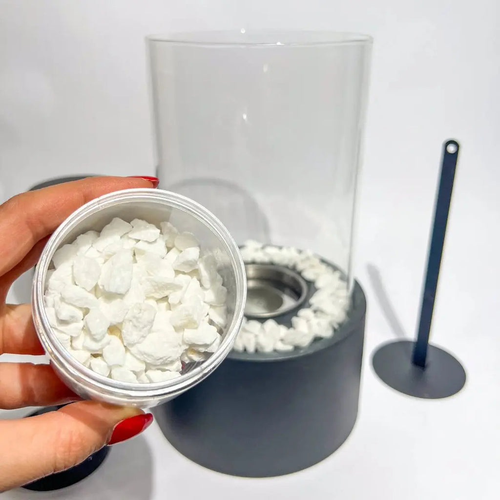 White Thassos marble gravel in hand beside a Helios tabletop fireplace with a metal tool on a white background.